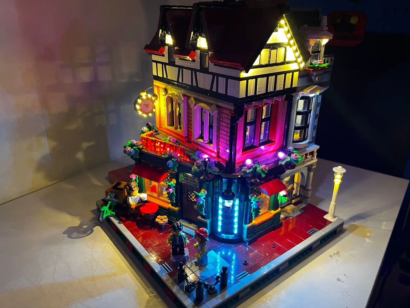 Impressive LEGO Icons Tudor Corner (10350) with 3266 pieces featuring custom LED lighting system. This British Tudor-style modular building showcases warm illumination throughout its multi-level structure including the ground floor restaurant and haberdashery, first-floor clockmaker's workshop, and cozy attic apartment. The half-timbered facade with steep red roof and rustic chimneys is beautifully highlighted by the lighting system. Standing 31cm tall by 26cm wide, this corner building includes 8 minifigures and connects seamlessly with other modular buildings in the collection.