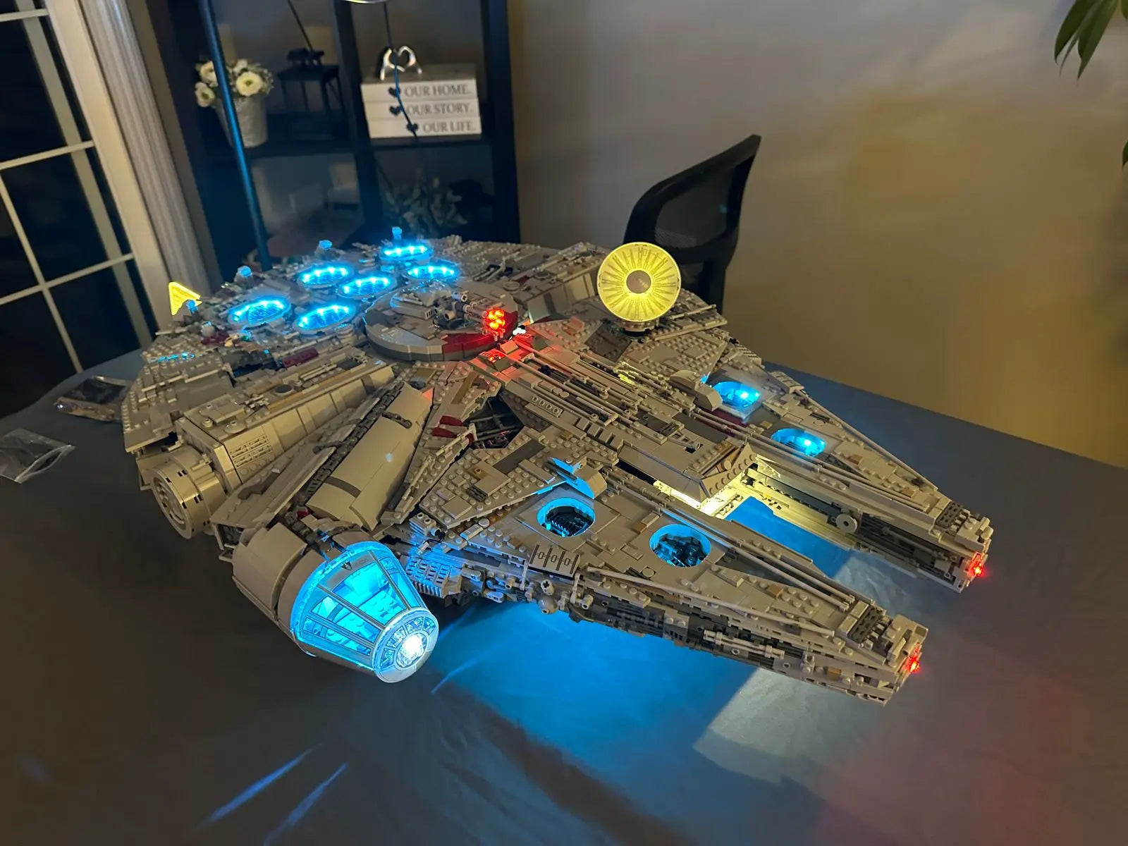 Spectacular LEGO Star Wars Millennium Falcon Ultimate Collector Series (75192) with 7500 pieces featuring custom LED lighting system. The legendary Corellian freighter displays blue illumination in the engines, cockpit, and various technical sections. Laser cannons are lit in red while a luminous yellow disc adorns the upper structure, recreating the authentic aesthetic of Han Solo's ship in a warm domestic setting.