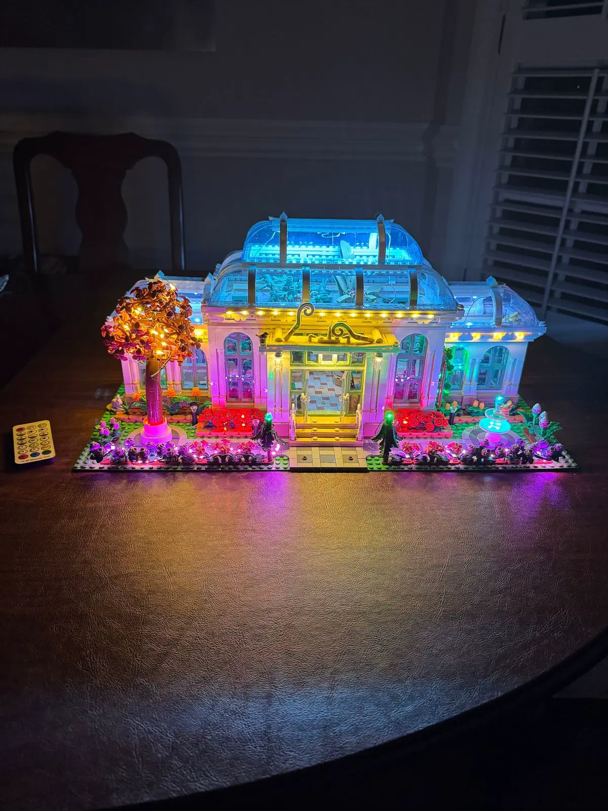 Magnificent LEGO Ideas Botanical Garden (21353) with 3792 pieces featuring custom LED lighting system. The Victorian-style glasshouse glows brilliantly with blue illumination in the upper conservatory sections, warm yellow lighting in the central entrance, vibrant pink and purple lights throughout the structure, and colorful ground lighting highlighting the exterior garden beds. The transparent walls and roof showcase over 35 plant species in this stunning botanical display model.